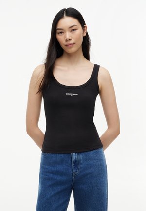Młoda kobieta z długimi czarnymi włosami, ubrana w czarny top z logo "tommy jeans" i niebieskie dżinsy, stoi na białym tle.