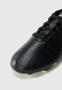 Detailný záber na čierne futbalové kopačky so zelenými diagonálnymi pruhmi, čiernymi šnúrkami a neonovo žltými kolíkmi na bielom pozadí.
