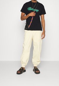 Czarna koszulka z zielonym grafikiem, kremowe dopasowane spodnie, czarne klapki z złotymi akcentami oraz mała torba na ramię z kolorowym paskiem.