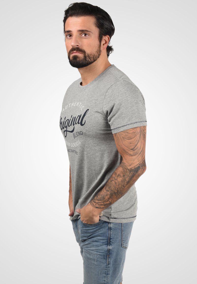 Pelēks īsā piedurkne t-krekls ar "Autentisks oriģināls" grafiku, ribināta kakla izgriezums un izliekta apakšmala, valkāts ar gaiši ziliem džinsiem.