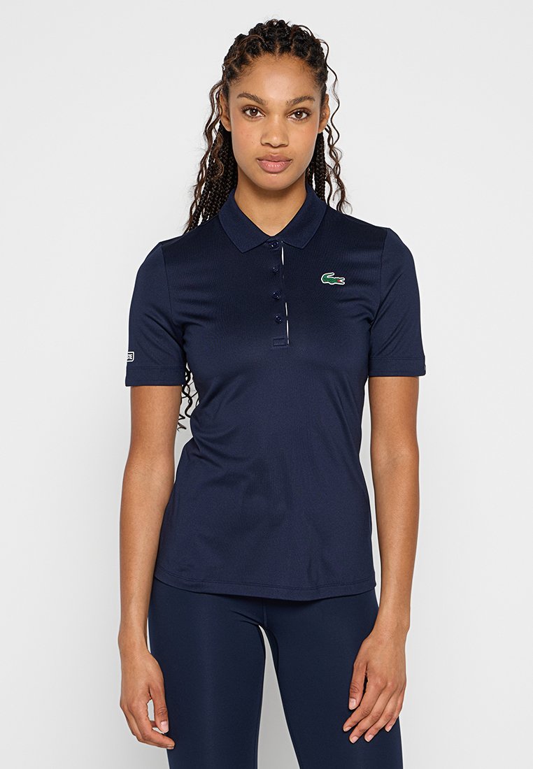 Granatowa koszulka polo wykonana z gładkiego materiału, z kołnierzykiem, krótkimi rękawami, zapięciem na trzy guziki oraz zielonym logo Lacoste.