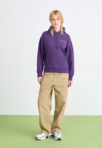 Lilas huppari, jossa on etutasku ja "Carhartt WIP" -logo. Yhdistetty vaaleanbeigeihin leveälahkeisiin housuihin ja valkoisiin tennoihin.