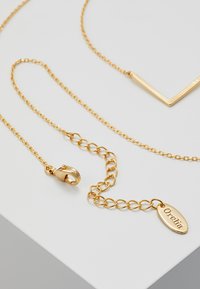 Goldkette mit einem V-förmigen Anhänger, Karabinerverschluss und einem ovalen Tag, auf dem "Orelia" eingraviert ist. Strukturierte Glieder und glänzende Oberfläche.