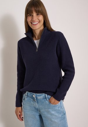 Femme souriante portant un pull zippé côtelé bleu marine et un jean bleu clair, une main dans la poche, sur un fond uni.