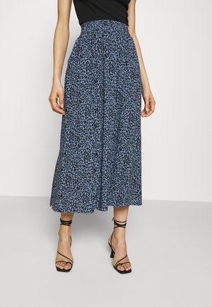 Femme portant une jupe midi taille haute à imprimé léopard bleu et noir, avec des talons noirs à lanières et un haut noir sans manches.