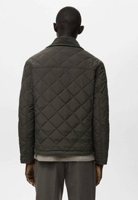 Giacca trapuntata verde scuro con collo rialzato e chiusura frontale con bottoni. Presenta cuciture diagonali e tessuto strutturato. Design pulito e minimale.