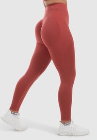 Leggings taille haute, ajustés, de couleur rouge rouille, fabriqués en tissu extensible, avec des coutures lisses et un léger éclat. À porter avec des baskets blanches.