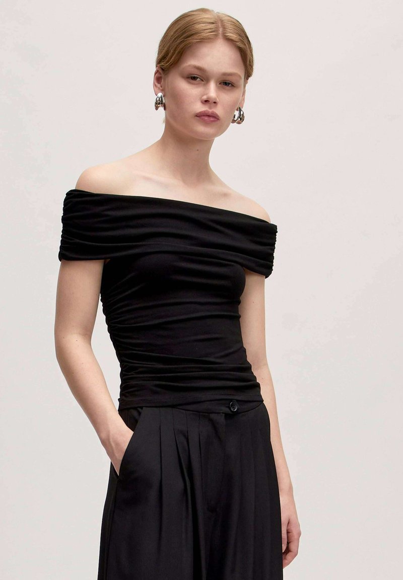 Zwarte off-shoulder top met gerimpelde textuur, aansluitend ontwerp en gladde stof. Gecombineerd met zwarte geplooide broek en knoopdetails.