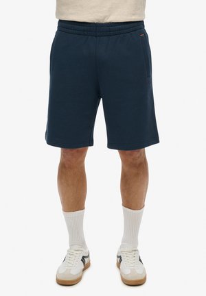 Pantalones cortos de algodón azul marino con cintura elástica, bolsillos laterales y un detalle de logo sutil. Combinados con calcetines blancos y zapatillas.