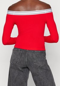 Henkilö, jolla on päällään punainen off-shoulder pitkähiha toppi, jossa on valkoiset raidat, sekä korkeavyötäröiset haalistuneet mustat farkut, kuvattuna selkäpuolelta.