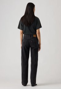 Γυναίκα με μακριά σκούρα μαλλιά, φοράει μαύρο κοντομάνικο πουκάμισο και φαρδιά ψηλόμεσα μαύρα τζιν, στέκεται αντικρίζοντας ένα απλό φωτεινό γκρι φόντο.