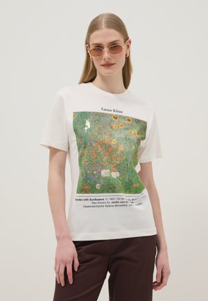 Femme aux cheveux blonds lisses portant des lunettes de soleil rondes et un t-shirt blanc imprimé du "Jardin aux tournesols" de Gustav Klimt.