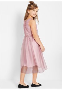 Robe sans manches rose pâle avec une jupe en tulle superposée et un dos en forme de trou de serrure, associée à des ballerines noires.