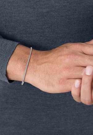 Silver metal bangle bracelet worn on a wrist, featuring a smooth, minimalist design and a subtle texture, contrasting against a dark sweater.