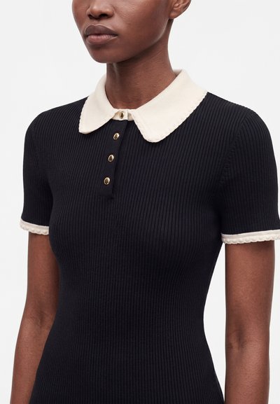 Femme portant un haut noir côtelé à manches courtes ajusté avec un col blanc festonné et des bords de manches assortis, et quatre boutons dorés.