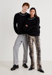 Dos personas están de pie una al lado de la otra. Una lleva un sudadera negra con texto blanco y jeans grises; la otra lleva una sudadera negra a juego y pantalones con un estampado.