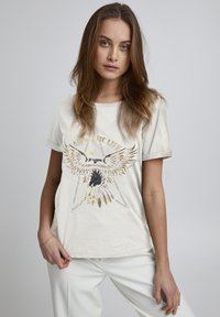 Kurzärmeliges cremefarbenes T-Shirt mit einem goldenen und schwarzen Adler-Design, versehen mit dem Text "AWAKENING LIFE" und einem lässigen Schnitt.