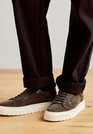 Baskets en daim marron avec semelles blanches épaisses et fermetures éclair latérales portées avec un pantalon noir retroussé sur un sol en bois.