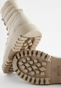 Beige gevoerde laars met een getextureerde rubberen zool met hexagonale patronen en richels. Voorzien van een trekriem voor gemakkelijk aantrekken.