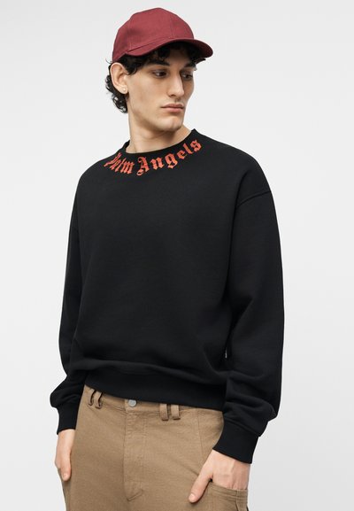 Νεαρός άνδρας που φοράει μαύρο φούτερ με την επιγραφή "Palm Angels" σε κόκκινα γοτθικά γράμματα στον γιακά, κόκκινο καπέλο και καφέ παντελόνι, κοιτάζει προς το πλάι.