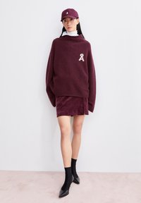 Burgunderfarbener Strickpullover mit strukturiertem Finish, versehen mit einem bestickten Logo. Kombiniert mit einem passenden Plüschrock und schwarzen spitzen Ballerinas.