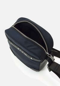Borsa a tracolla in nylon blu navy con zip argentata, dettagli neri e una placca con logo. Presenta una superficie liscia e una tracolla regolabile.