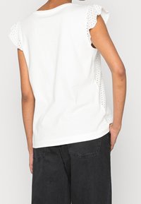 Witte katoenen top met eyelet-stof zijpanelen en geruite mouwen. Relaxte pasvorm en gebogen zoom. Gecombineerd met donkere spijkerbroeken.