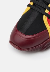 Gros plan sur l’avant d’une chaussure de sport en cuir grenat, tissu noir, bordure jaune et lacets noirs sur fond blanc.