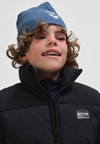 Zēns, kurš valkā melnu polsterētu jaku ar augstu apkakli un zilu cepurīti ar baltu logotipu, redzami viļņaini mati un viegls smaids.