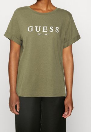 Personne portant un t-shirt manches courtes vert olive avec des revers sur les manches et un texte blanc "GUESS EST. 1981" sur le devant.