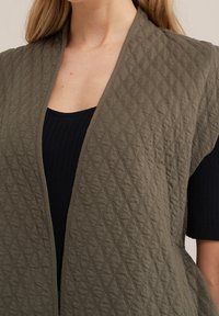 Oliivinvihreä tikattu liivi, jossa timanttikuvio, edestä avoin ja teksturoitu kangas. Päällä mustan ribbineuletakin päällä.