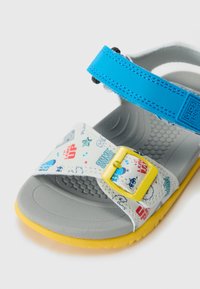 Grå sandal med et mønstrete stropp som har blå og gule aksenter, justerbar blå borrelås og en teksturert gul såle.