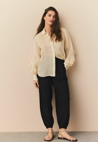 Lys beige, semi-gennemsigtig bluse med volanteærmer, parret med løstsiddende sorte bukser og gyldne flade sandaler. Simpelt, elegant design.