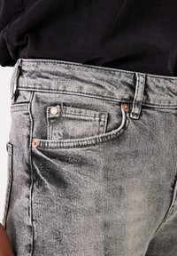 Vaqueros de mezclilla gris con una textura desgastada, diseño de cinco bolsillos y acentos de hardware en tono cobre a lo largo de la cintura y los bolsillos.