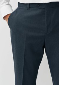 Navy trousers made of textured fabric with a fine pattern, featuring a flat front, side pockets, and a single button closure at the waist.