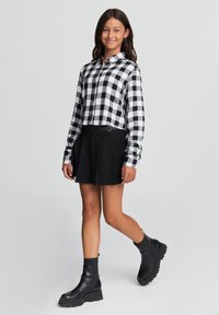 Giovane che cammina, indossando una camicia a scacchi bianca e nera, una gonna plissettata nera e anfibi neri spessi, su uno sfondo chiaro e semplice.