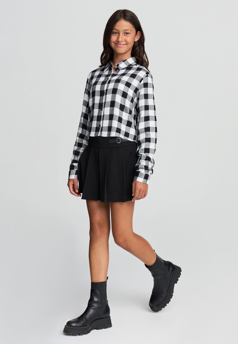 Giovane che cammina, indossando una camicia a scacchi bianca e nera, una gonna plissettata nera e anfibi neri spessi, su uno sfondo chiaro e semplice.