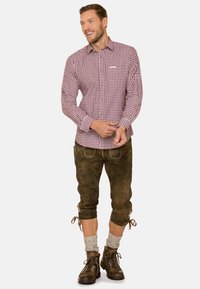 Ein rot-weiß kariertes Button-Down-Hemd kombiniert mit braunen Lederhosen in Knielänge. Dazu werden graue Socken und braune Schnürstiefel getragen.