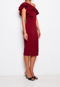 Burgunderfarbenes schulterfreies Kleid mit Rüschendetails, figurbetontem Schnitt und knielanger Saumlänge. Das Model trägt schwarze Sandalen mit offenen Zehen.