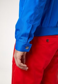 Modrá nylonová bomber bunda s elastickými manžetami a kovovým knoflíkem, kombinovaná s červenými kalhotami. Zaměření na texturu a barevný kontrast.