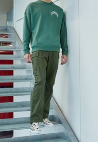 Zelená mikina s logem "Champion", spárovaná s olivovými cargo kalhotami a černobílými teniskami s potiskem, stojící na schodišti.