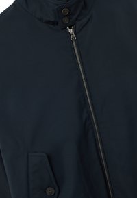 Giacca blu scuro con una cerniera diagonale argentata, fascetta del colletto con bottone e tasca con flap e bottone sul davanti.