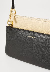Borsa a polso in pelle bicolore, nera e beige, con zip dorate, anelli e catena, caratterizzata dal nome del marchio COCCINELLE sul davanti.