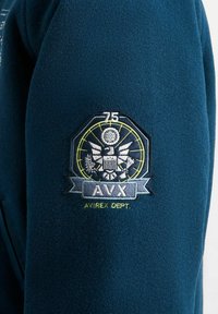Blauwe stof met een geborduurd patch met een adelaar, "75" erboven, "AVX" in een banner en de tekst "AVIREX DEPT." eronder.