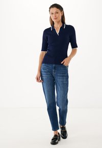 Granatowa koszulka polo z krótkim rękawem, z białymi wykończeniami i falbaniastym dołem, w zestawie z jasnoniebieskimi spodniami o prostym kroju i czarnym obuwiem.