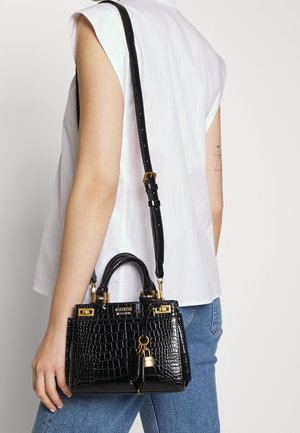 Personne portant un haut blanc sans manches et un jean bleu tient un sac à main Guess noir embossé effet croco avec des éléments dorés sur l'épaule.
