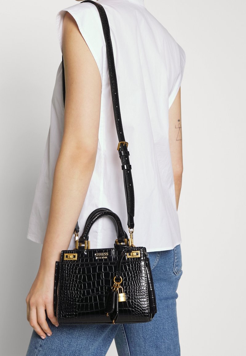 Personne portant un haut blanc sans manches et un jean bleu tient un sac à main Guess noir embossé effet croco avec des éléments dorés sur l'épaule.