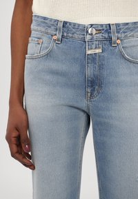 Jean en denim bleu clair avec un design classique à cinq poches, comprenant des accessoires en ton cuivre et un léger effet délavé sur le tissu.