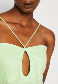 Hellgrünes ärmelloses Kleid mit einem Schlüsselgraben-Ausschnitt und dünnen Trägern, aus strukturiertem Stoff. Klare Linien und sanfte Kanten.