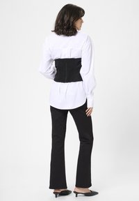 Sort smocket korsett over en hvid skjorte, båret med sorte flared bukser og sorte hælede mules. Glat stof med struktureret design.